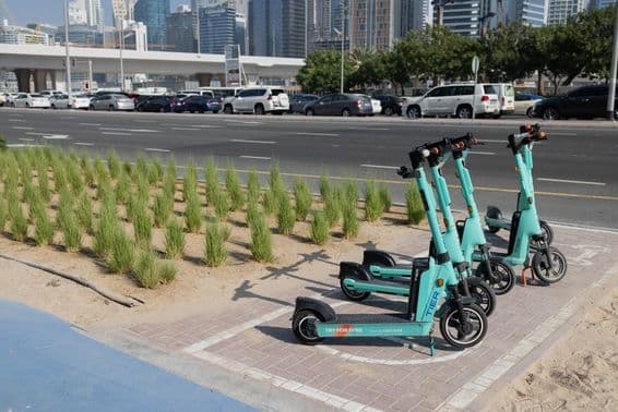 Trottinettes électriques garées devant un gratte-ciel de Dubaï avec des plantes vertes sur le séparateur de route.