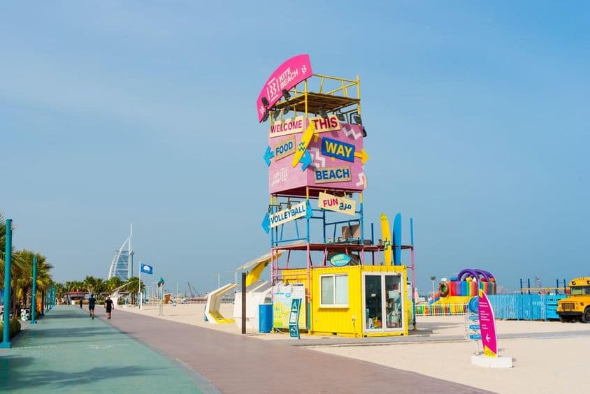 Structure d'accueil de Dubai Kite Beach, panneaux, Burj Al Arab en fond, premier plan avec piste de course et plage de sable.