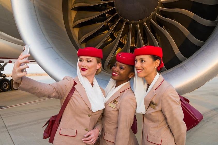 Trois hôtesses de l'air Emirates prenant un selfie devant un moteur d'avion Emirates.