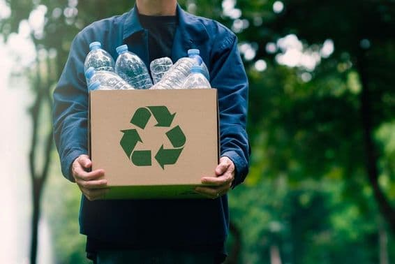 Une personne portant un carton avec des bouteilles consignées, avec des arbres verts flous en arrière-plan.
