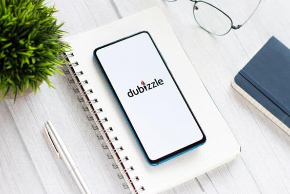 Un téléphone affichant Dubizzle sur une table, sous un bloc-notes, à côté de lunettes et d'une petite plante verte.