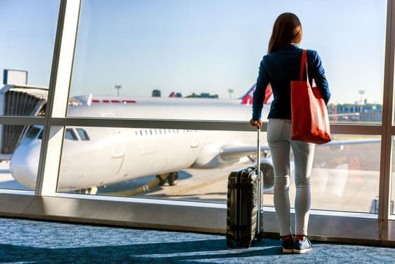 Une femme se tient à la fenêtre avec un avion visible, tenant une valise à roulettes de sa main gauche et un sac à main dans sa main droite.