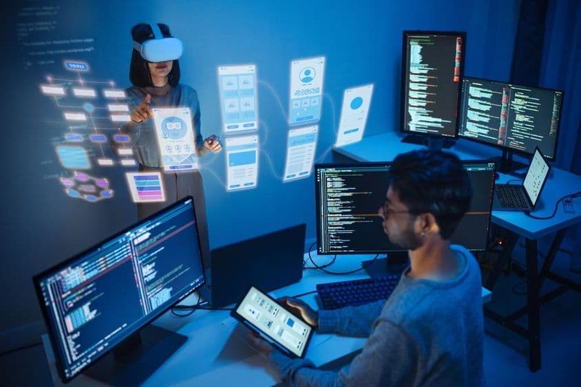 Un développeur d'applis assis devant un ordinateur, tenant une tablette, avec deux écrans affichant du code informatique.