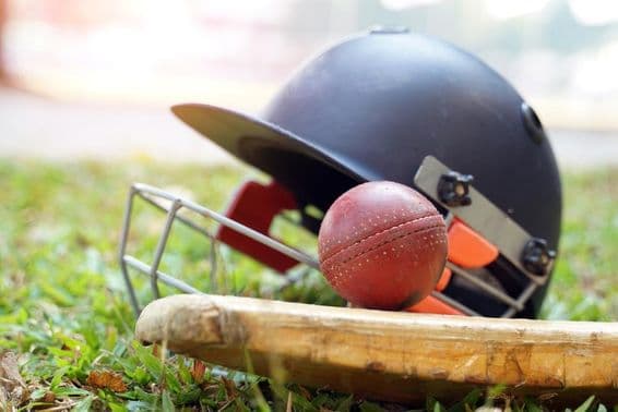 Une batte de cricket avec une balle au-dessus et derrière elle, un casque de cricket est visible sur un champ herbeux.