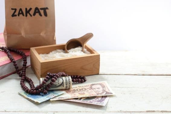 Sac étiqueté Zakat devant une boîte contenant du riz, des perles, des dollars et d'autres devises.