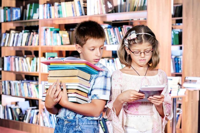 Deux enfants à la bibliothèque, un lisant un e-book, l'autre portant de nombreux livres dans ses mains.