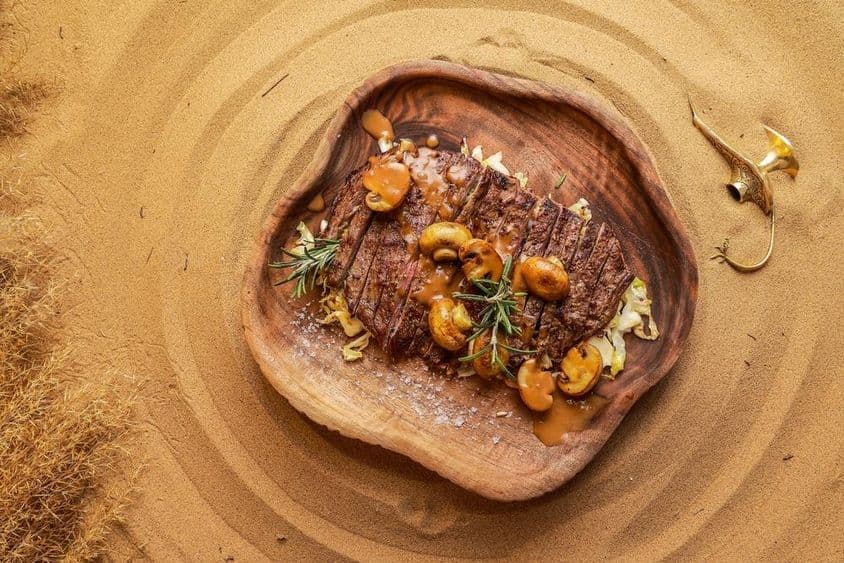 Steak servi avec des champignons sur une planche en bois dans le sable du désert, avec une lanterne éteinte à côté.