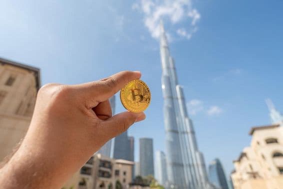 Personne tenant une pièce d'or Bitcoin, face au ciel, avec le Burj Khalifa en arrière-plan.