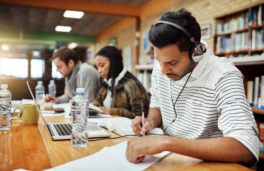 Étudiants universitaires écrivant sur papier avec livre et cahier devant, l'un portant des écouteurs.