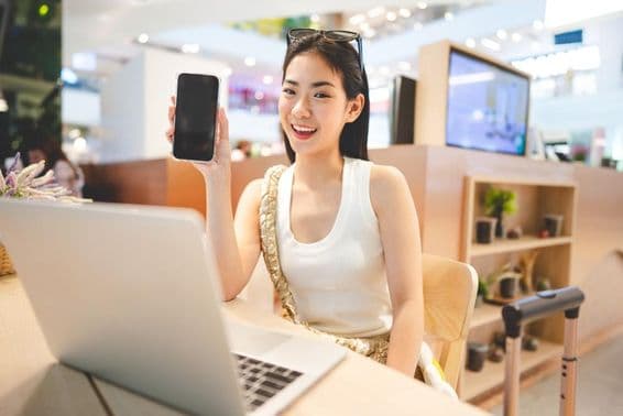 Fille de la Génération Z travaillant, tenant un téléphone, avec un cahier devant elle et une poignée de valise à roulettes à côté d'elle.