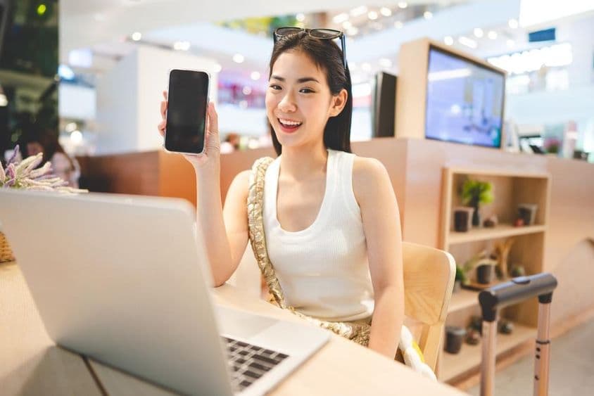 Fille de la Génération Z travaillant, tenant un téléphone, avec un cahier devant elle et une poignée de valise à roulettes à côté d'elle.