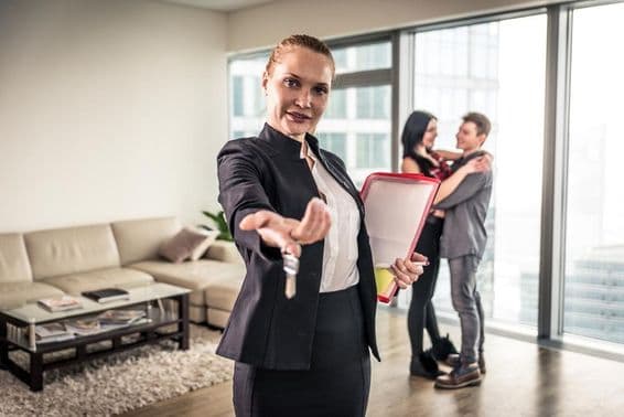 Une agente immobilière saluant de la main, un couple s'enlaçant en arrière-plan.