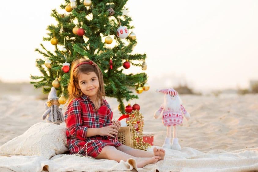Jolie petite fille souriante assise sur la plage sous un sapin de Noël.