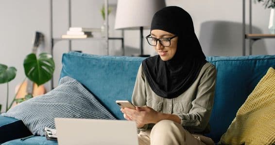 Fille arabe en hijab travaillant à domicile utilisant un smartphone et un ordinateur portable tout en étant assise sur le canapé.