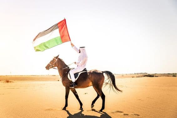 Homme du Moyen-Orient en tenue traditionnelle émiratie montant un cheval arabe dans le désert de Dubaï.