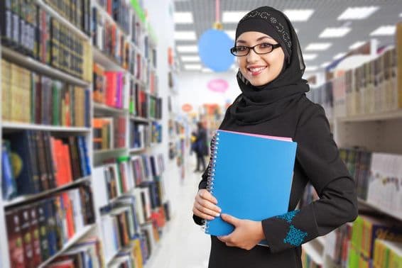 Étudiant arabe se tient dans la bibliothèque avec un cahier à la main.