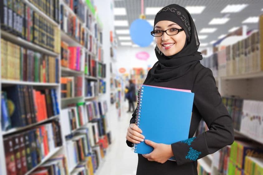 Étudiant arabe se tient dans la bibliothèque avec un cahier à la main.