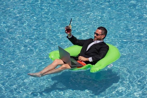 Homme d'affaires prospère flottant en costume dans l'eau de la piscine, buvant un cocktail d'été et utilisant un ordinateur portable dans la piscine.