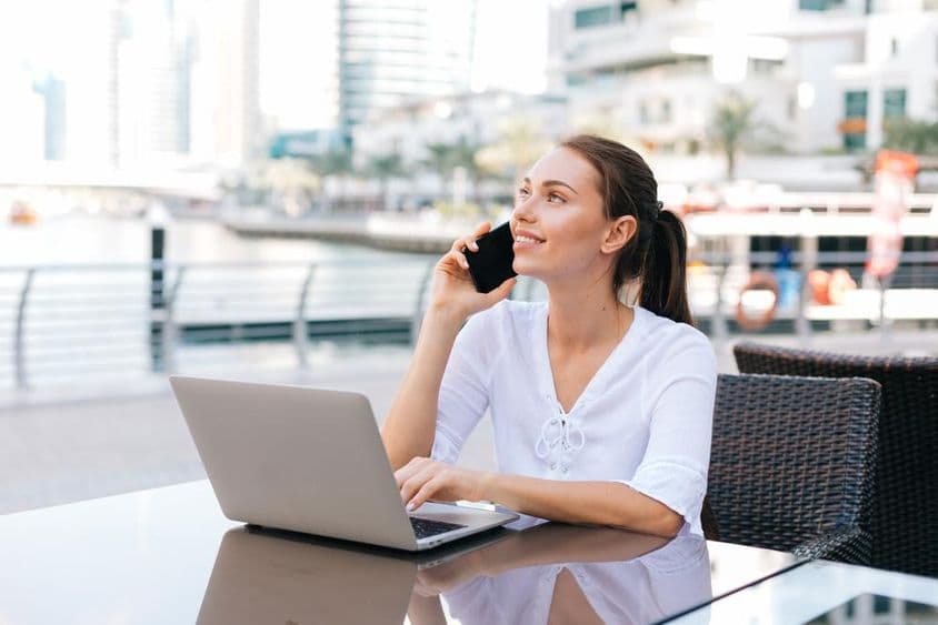 Jeune femme freelance travaillant sur un ordinateur portable, assise dans un café en plein air tenant un téléphone.