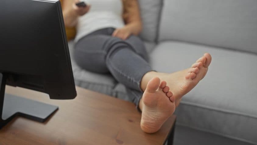 Une jeune femme allongée pieds nus sur un canapé confortable dans le salon, tenant une télécommande.