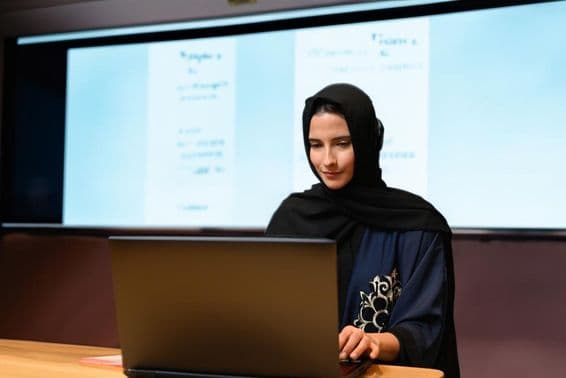 Enseignante portant un Abaya Hijab avec un écran de projecteur en arrière-plan dans une salle de classe moderne avec écran numérique.