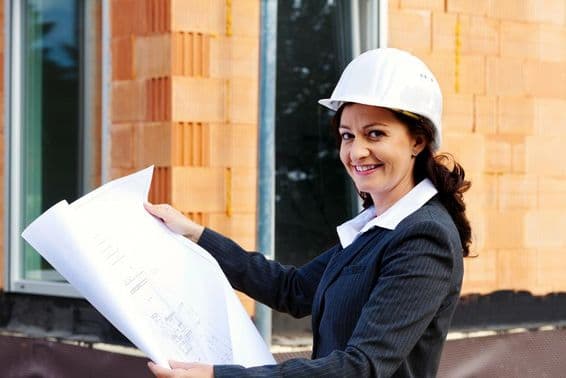 Femme architecte avec plan en main devant une maison en construction, portant un casque de sécurité.