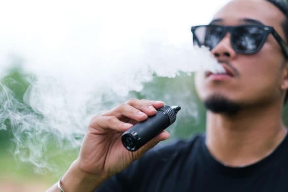 Homme asiatique fumant une cigarette électronique, expirant de la fumée par la bouche et profitant de l'extérieur en été.