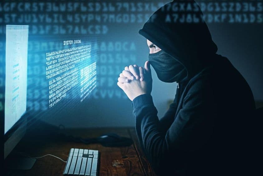 Cybercriminalité, homme encapuchonné assis devant un ordinateur.