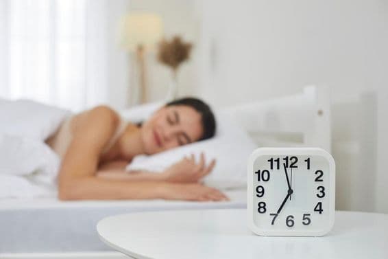 Réveil sur une table de chevet dans une chambre, avec une jeune femme dormant en arrière-plan.