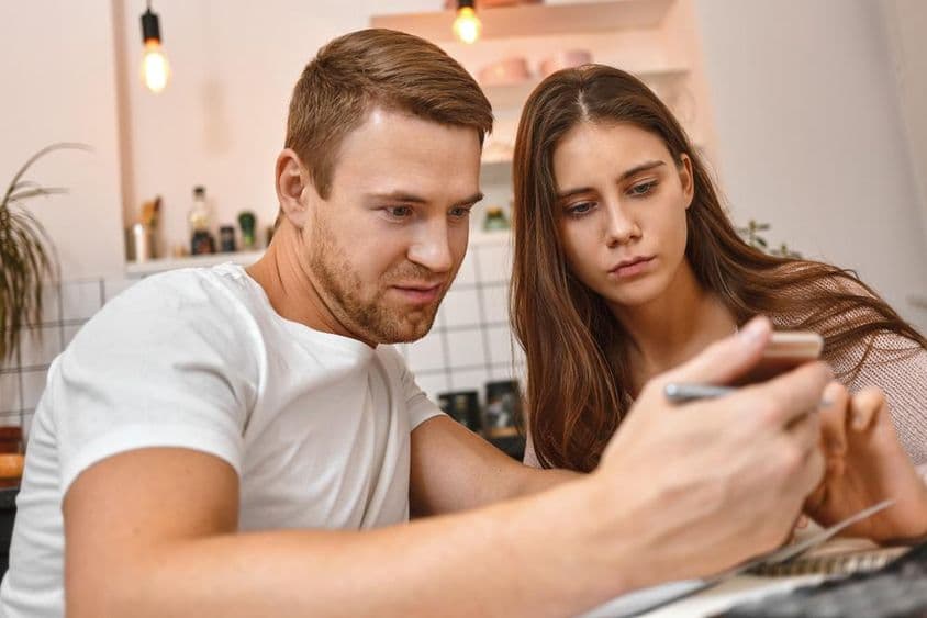 Jeune couple savourant un café dans la cuisine en lisant une notification bancaire en ligne sur un téléphone mobile.