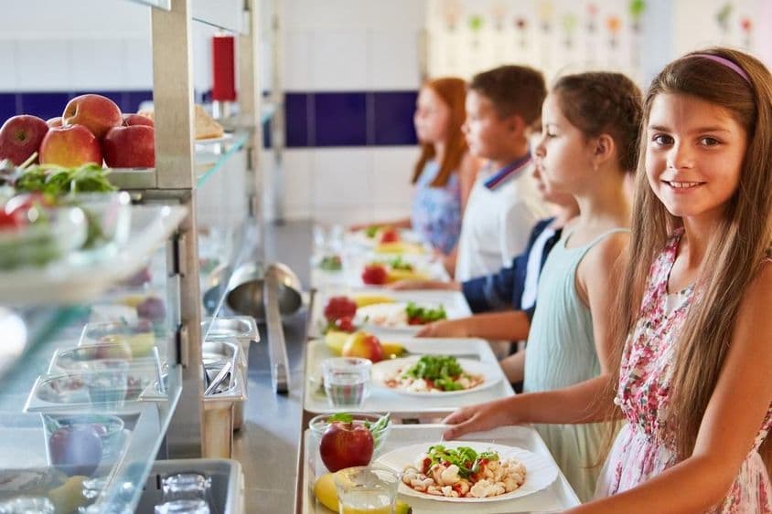 Fille souriante avec un plateau repas, en ligne avec des amis pendant la pause déjeuner à la cafétéria de l'école.