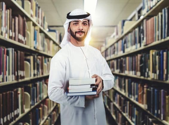 Étudiant arabe debout dans une bibliothèque tenant des livres.