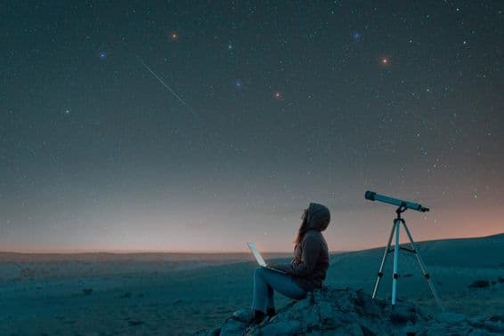 Femme assise dans le désert avec un ordinateur portable à côté d'un télescope la nuit, regardant le ciel étoilé.