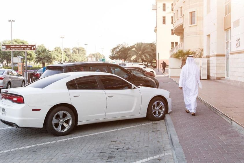 Dans les rues de Dubaï : Un homme en tenue nationale et une Dodge blanche dans le parking.