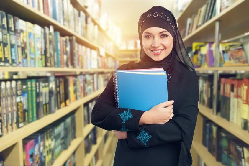 Étudiant arabe debout dans une bibliothèque avec des livres dans les bras.