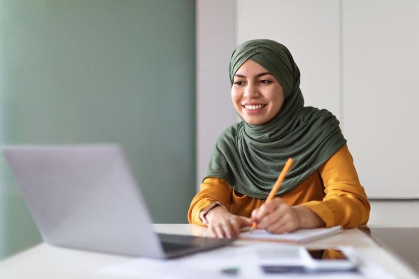 Jeune femme musulmane regardant un webinaire à domicile sur un ordinateur portable et prenant des notes.