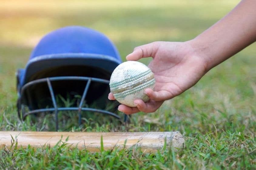 La main d'un joueur choisissant une balle de cricket parmi d'autres équipements de cricket posés sur l'herbe.