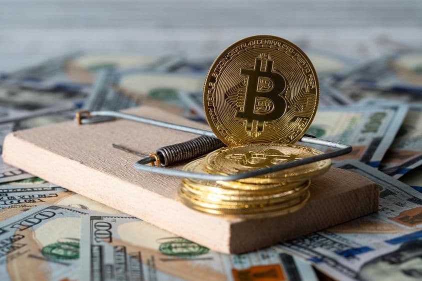 Pièces de cryptomonnaie en or bitcoin dans un piège à souris, avec des billets de cent dollars américains en arrière-plan.